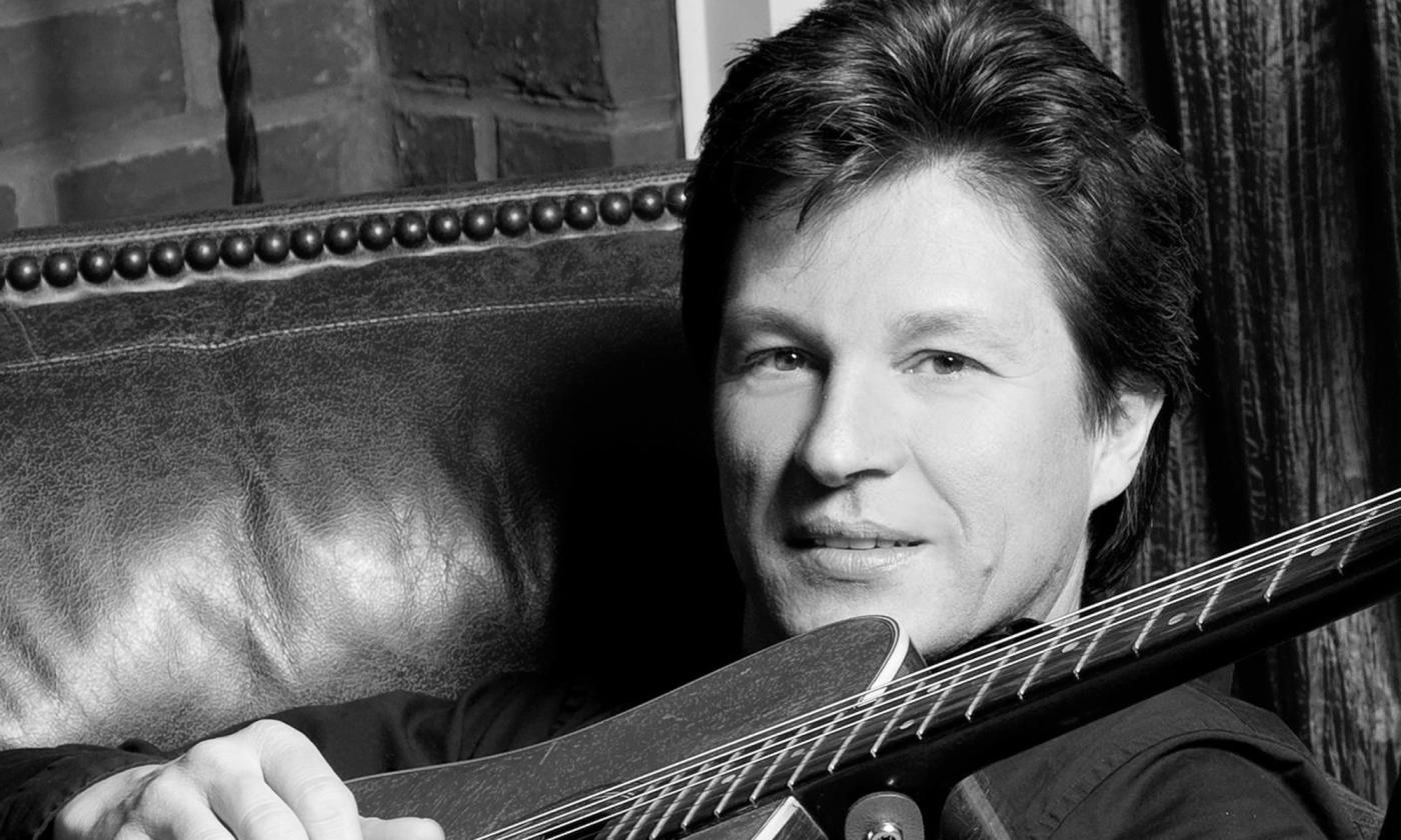 B&W Close up of a middle-aged white man sitting on a leather chair with a guitar propped across his body