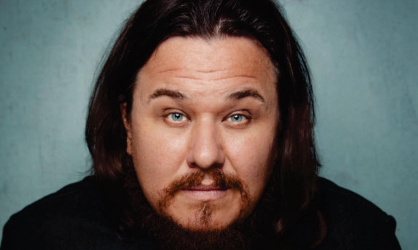 Shane Torres poses in black clothing and sits in front of a grey background.