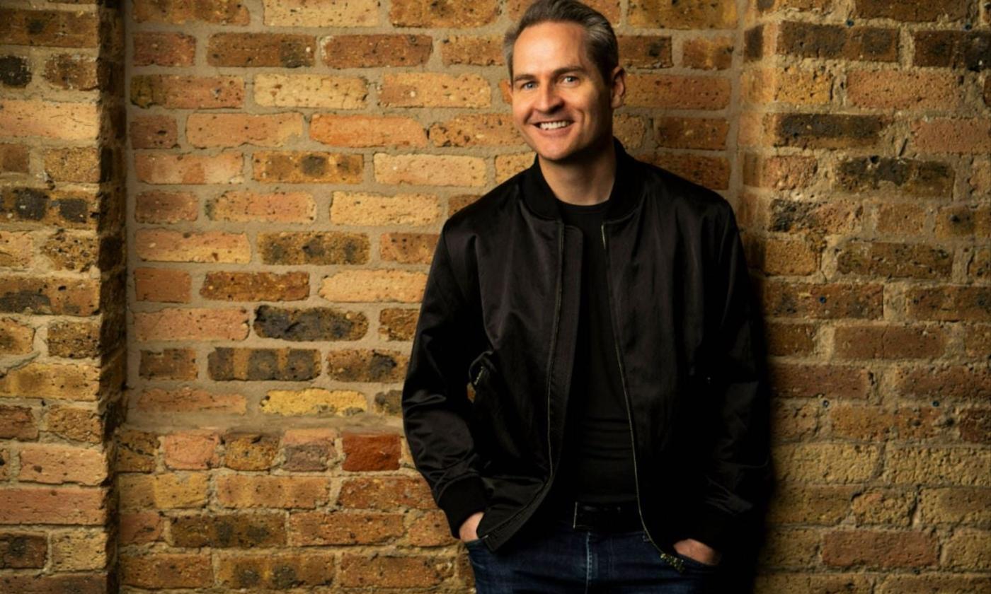 Pat McGann smiles and poses in a leather jacket in front of a brown brick wall.