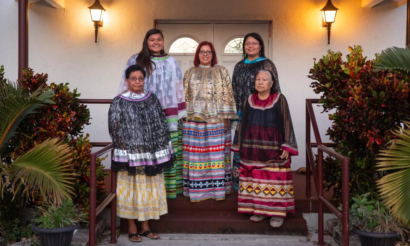 Five Native-American women in their tribal clothing, on the steps of a modern adobe building