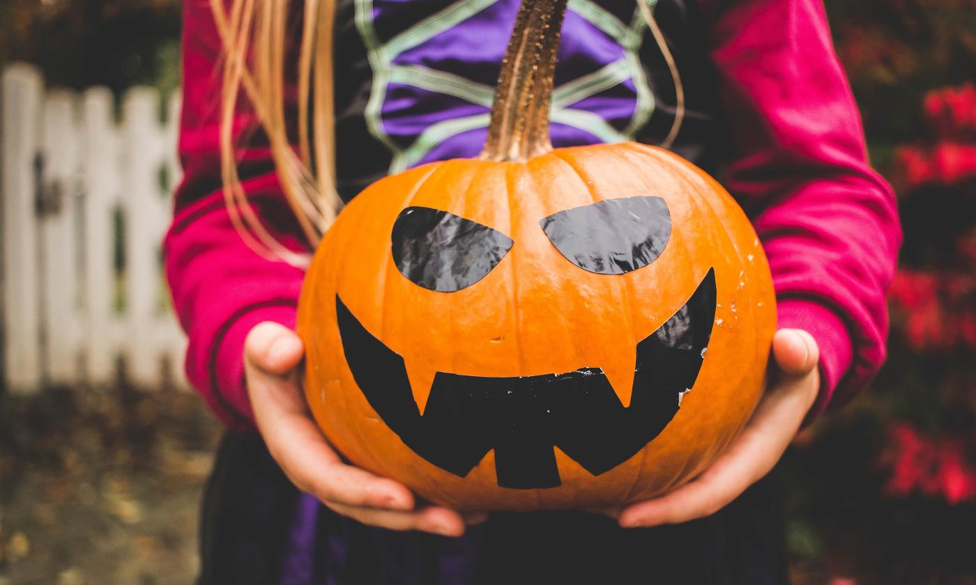 A pumpkin with a painted spooky face, being held in the arms of a costumed child