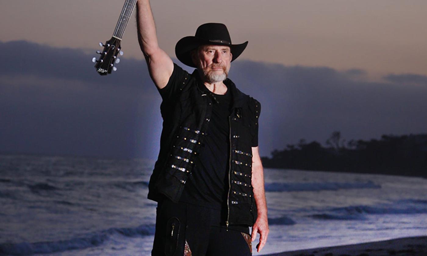 Colin Hay poses in black clothing while holding his guitar. 