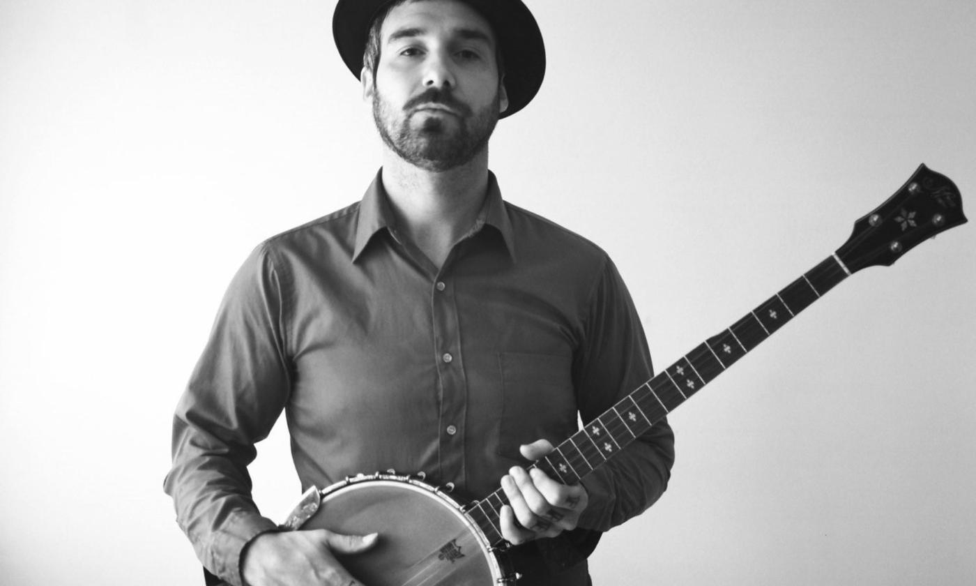 William Elliott Whitmore poses in a black and white photo while wearing a hat and holding his musical instrument.
