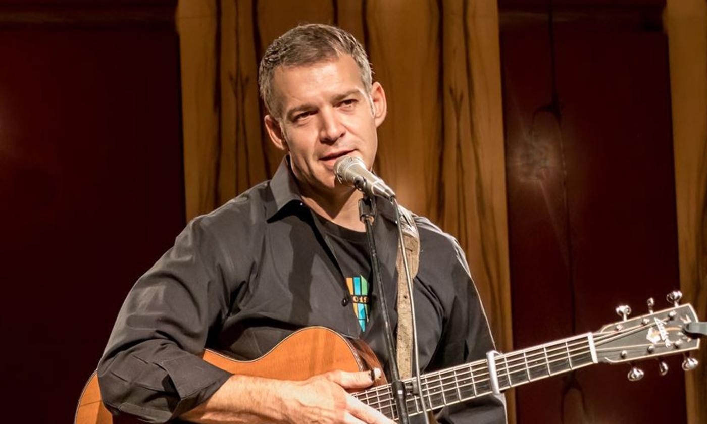 Sam Pacetti with guitar, speaking into microphone