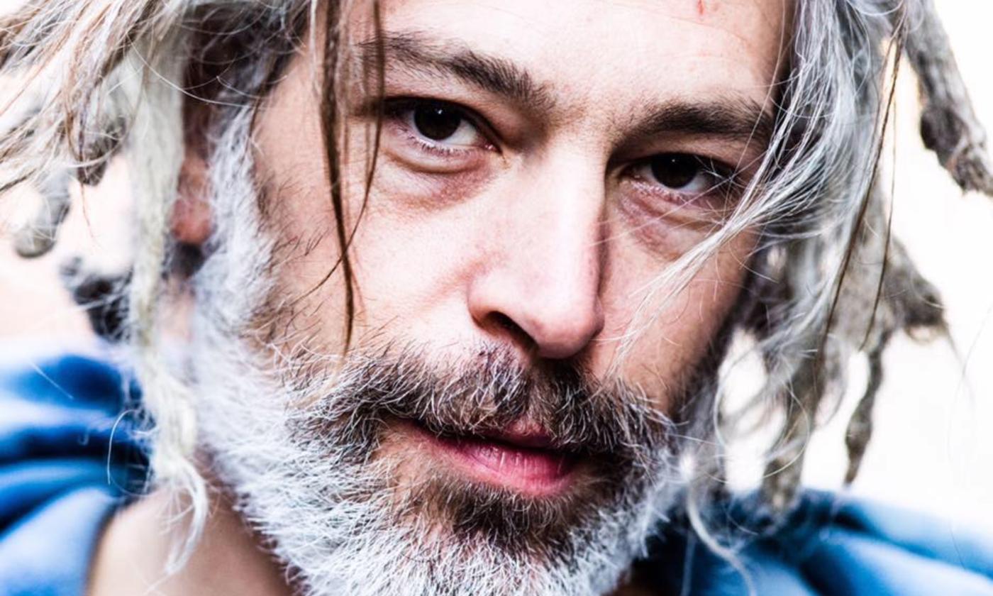 A headshot of Matisyahu wearing a blue shirt and posing in front of a white backdrop. 