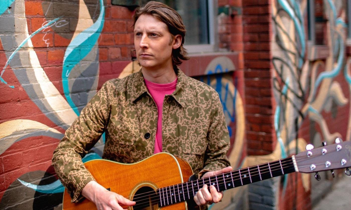 Eric Hutchinson poses with his guitar in front of a colorful brick wall. 