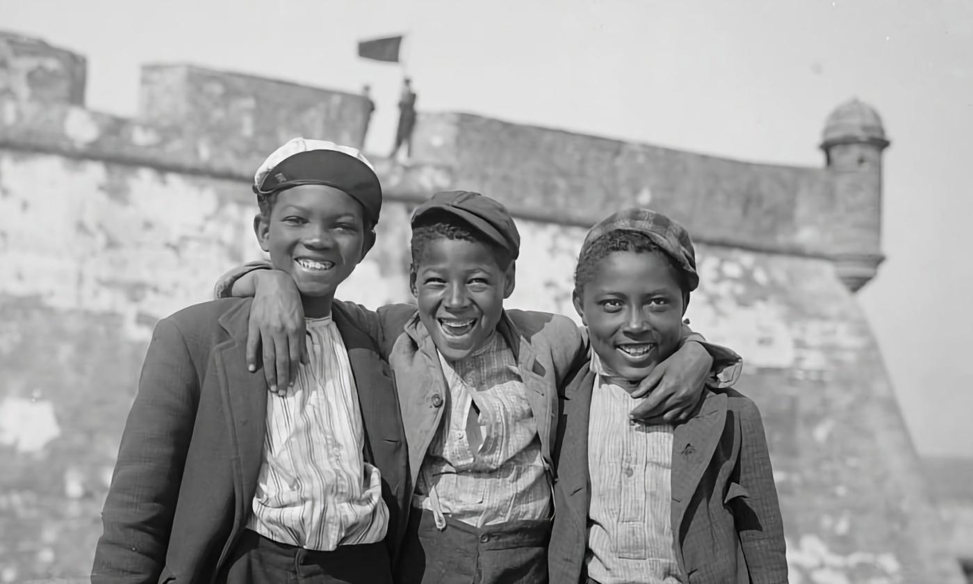 B&W circa 1890 Three young Black children smile and pose in front of the Castillo de San Marcos. The middle child has his arms around his two friends, also male.