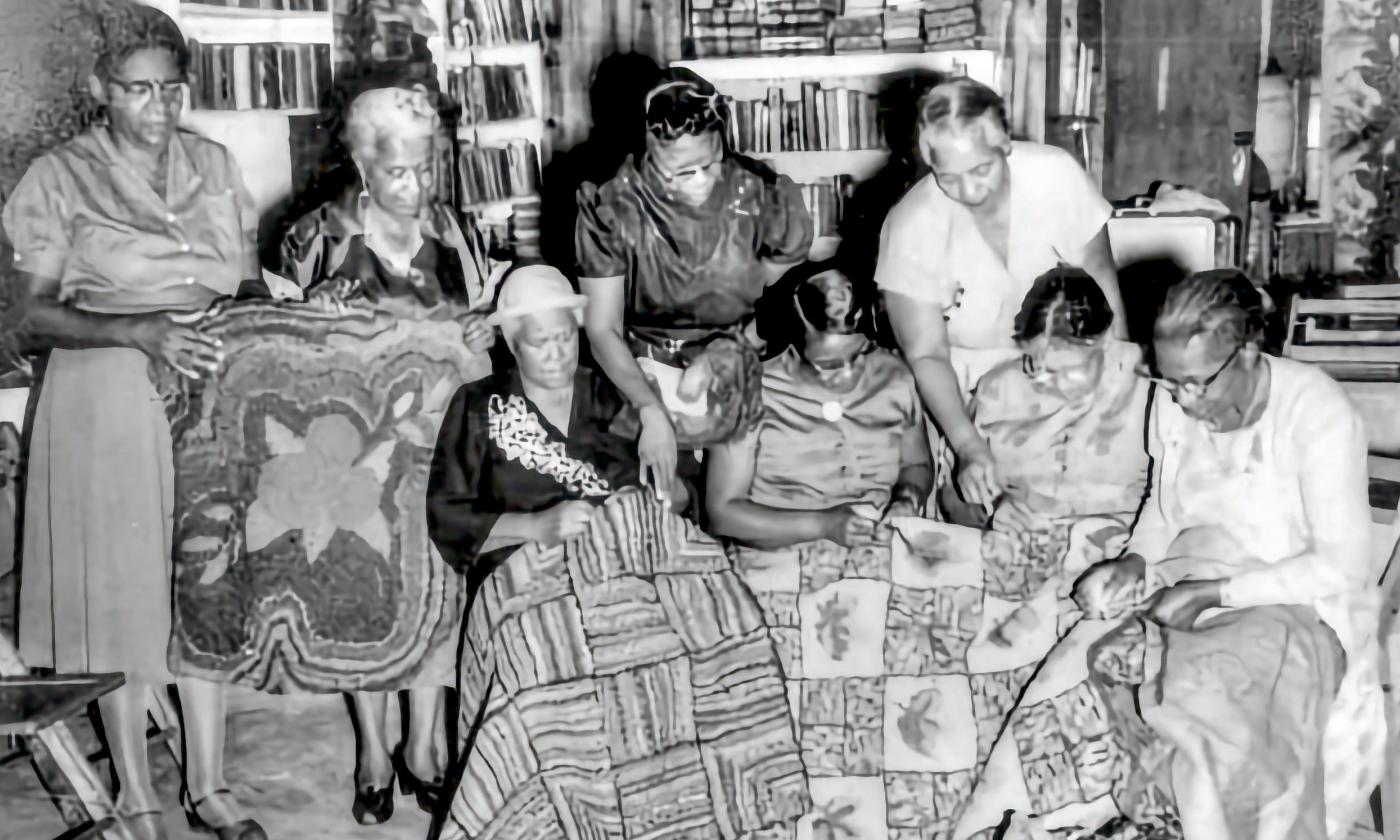 B&W a group of Black women display and discuss their quilts and crafts, which sit on their laps.