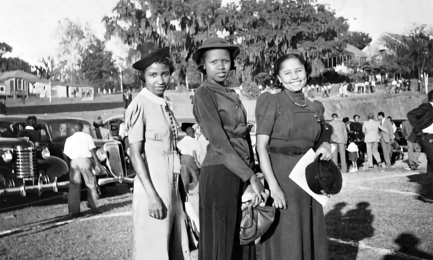 B&W, three young Black women pose in front of a line of cars.