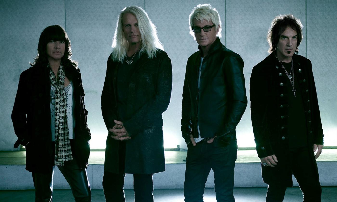 Bandmates from REO Speedwagon wear black clothing while posing in front of a well-lit backdrop.