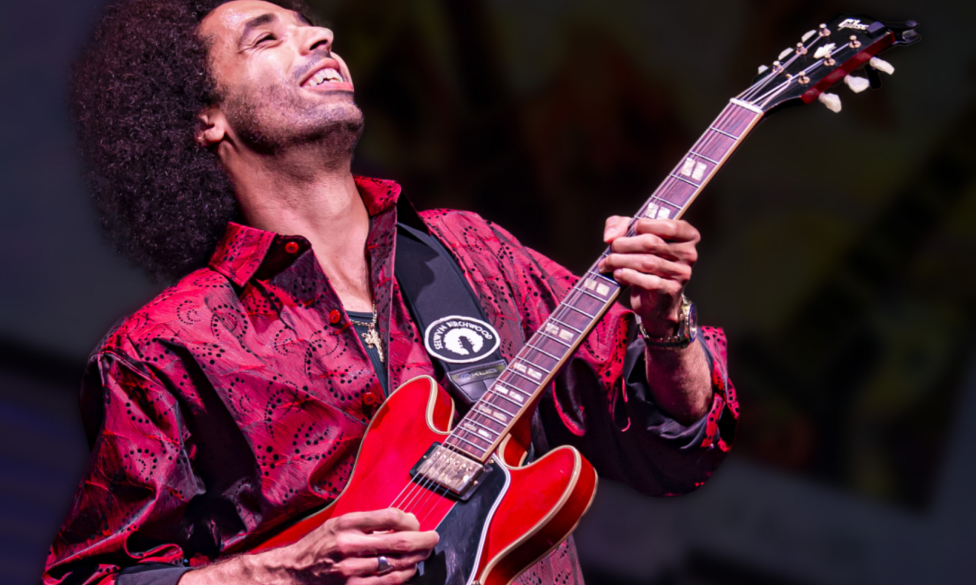 Selwyn Birchwood holds his guitar and smiles in front of a black backdrop. 