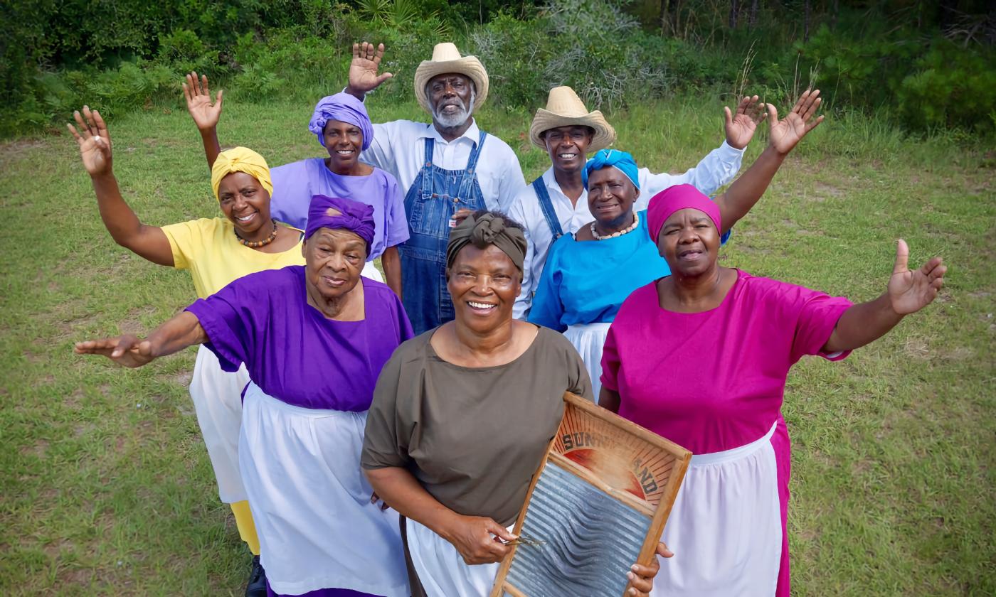 Eight members of the Gullah Gitchee Ring Shouters, standing on a lawn and waving