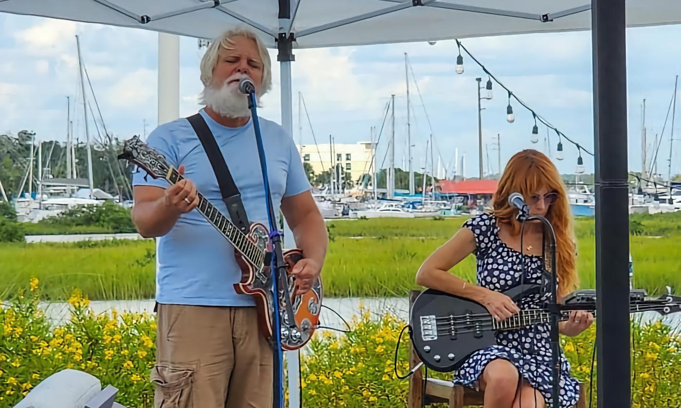 Bryson and Amy Hendricks performing at a venue on the banks of the San Sebastian River, with river and docked boats at their backs