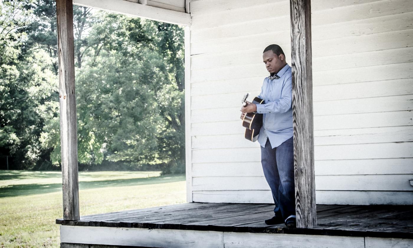 Jontavious Willis, standing on the porch of a white-sided building, playing guitar