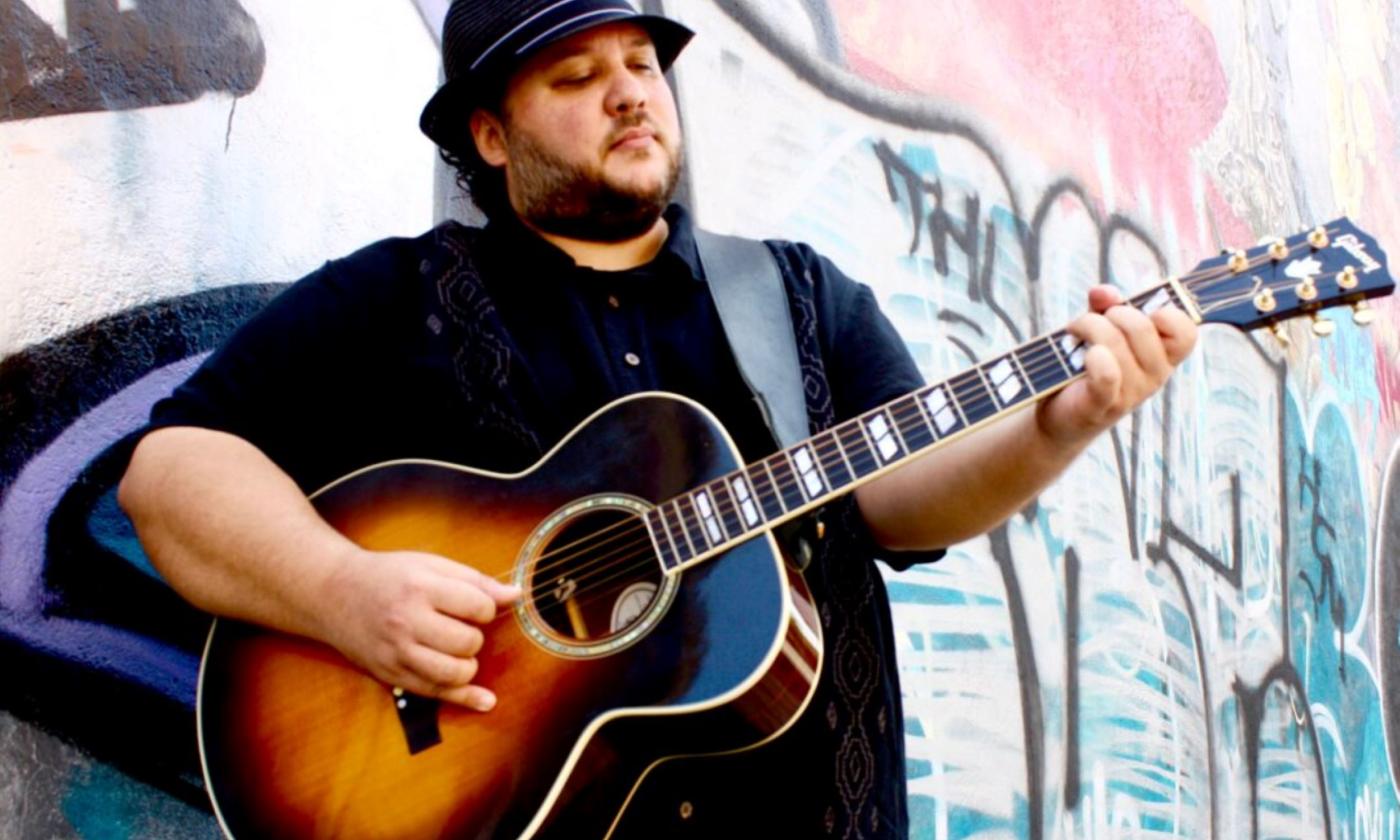 Mutlu clutches his guitar and poses in front of a wall with graffiti. 