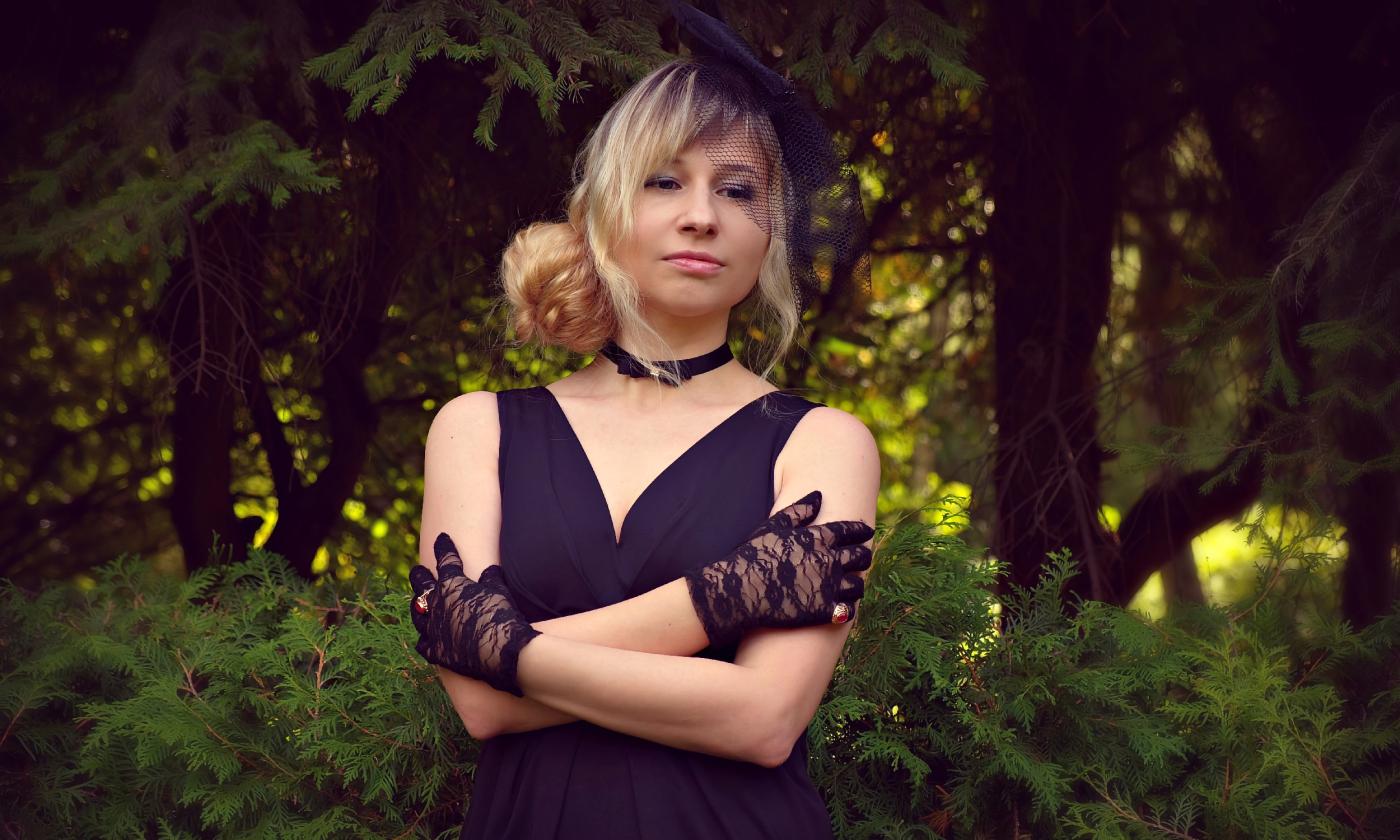 A blonde woman wearing a black dress, cloves, and fascinator, standing in a garden of trees