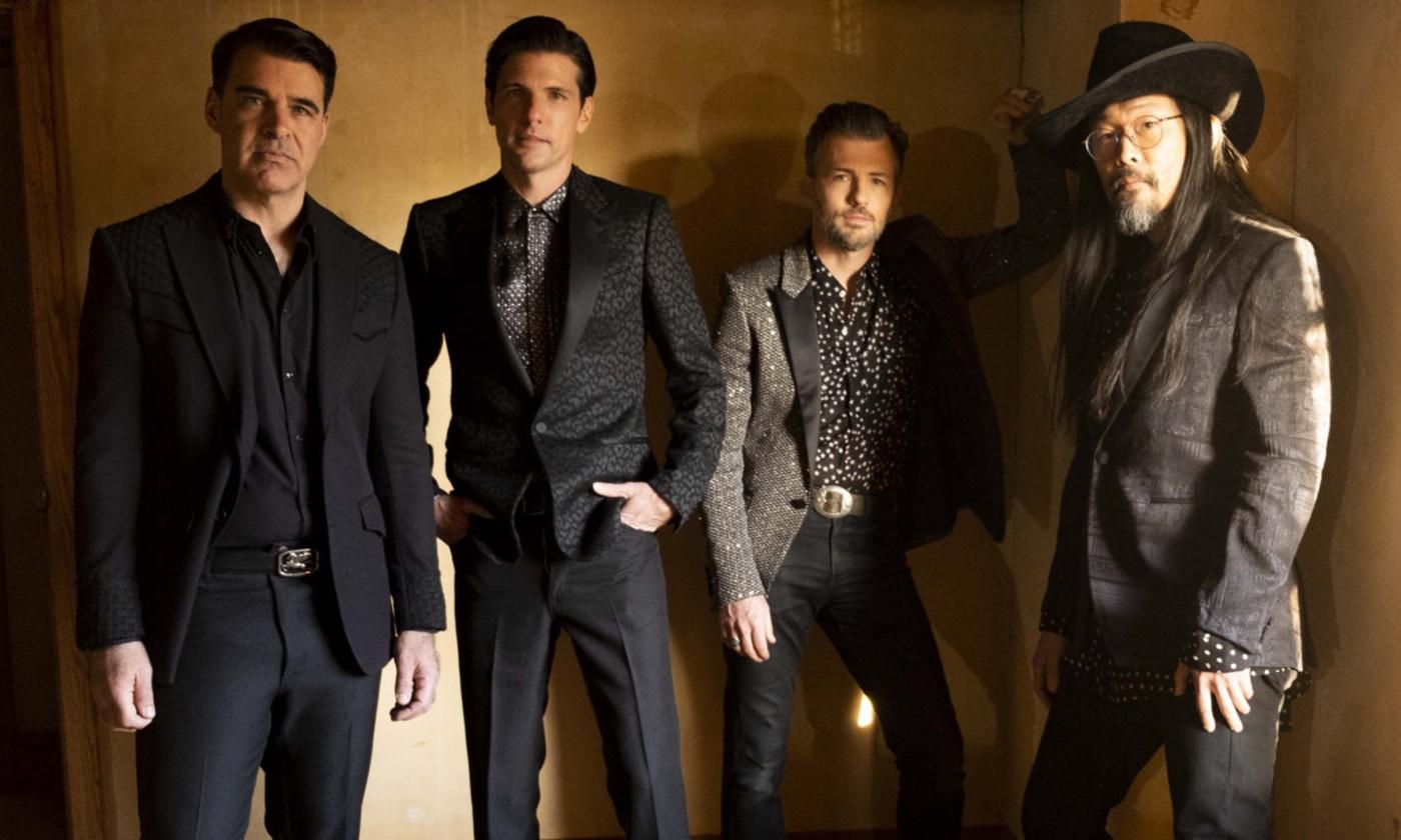 The Avett Brothers wear black suits and blazers while posing in front of a brown backdrop.