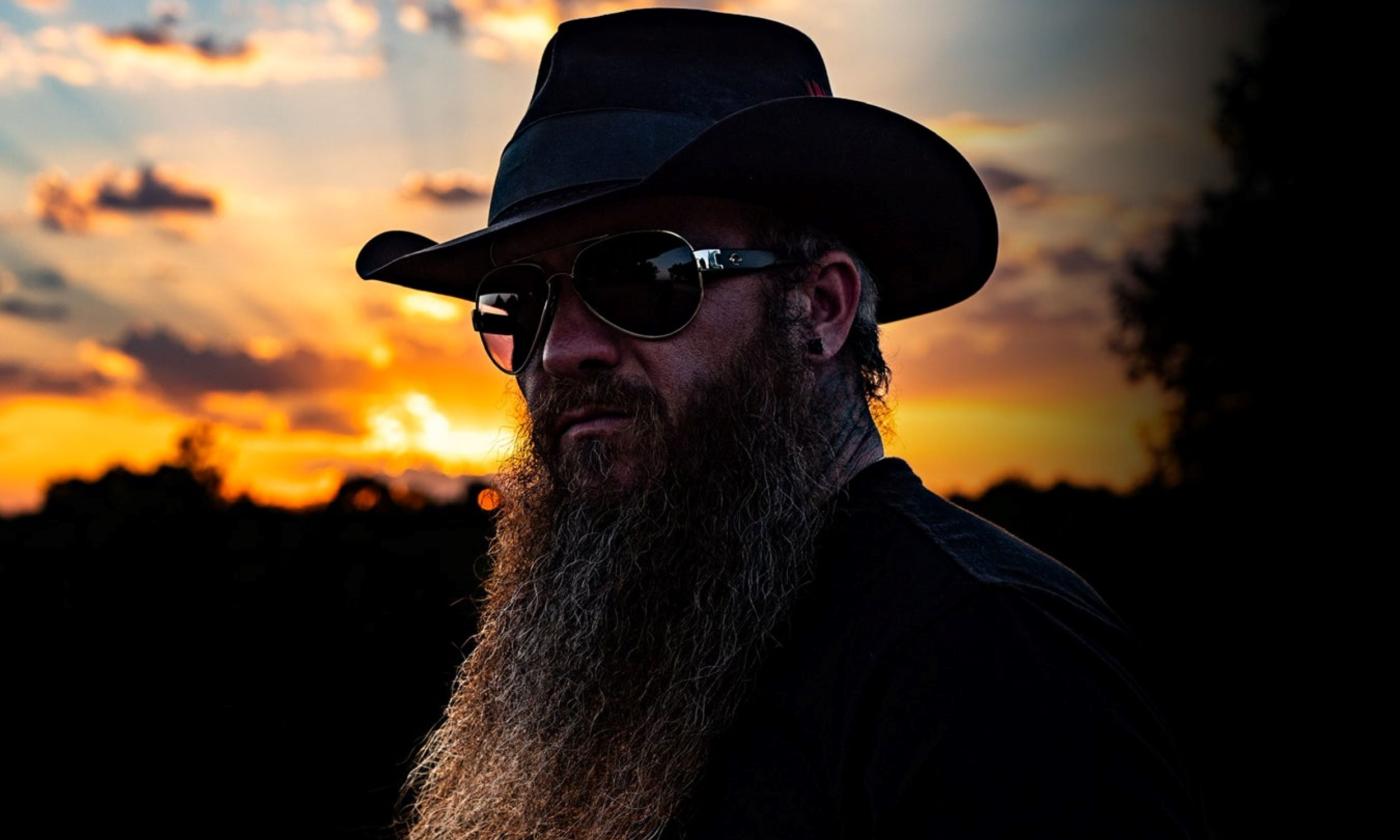 Cody Jinks wears a dark hat with matching sunglasses with the sunset as a backdrop.