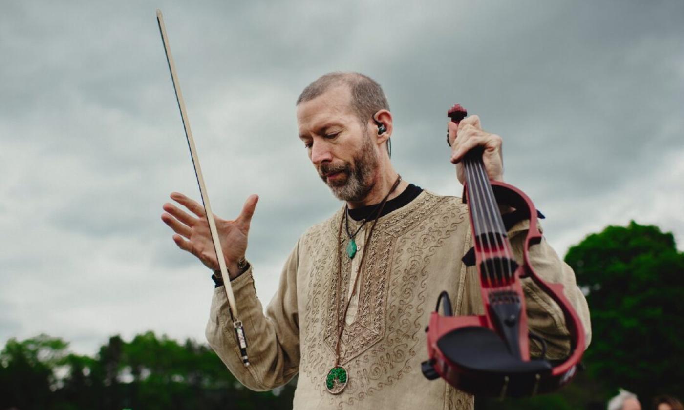 Dixon holds his violin and poses in front of a cluster of trees and bushes. 