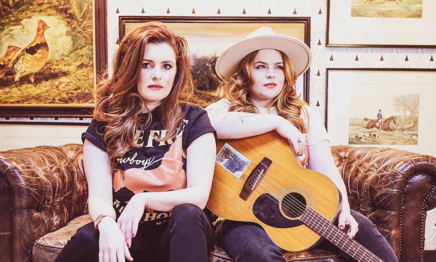 Cassie and Maggie pose side by side on a brown chesterfield couch in front of pictures set in a frame.