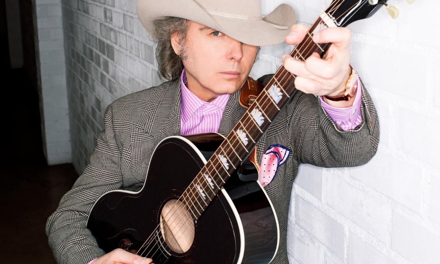 Dwight Yoakam strums his guitar and poses in front of a white wall.