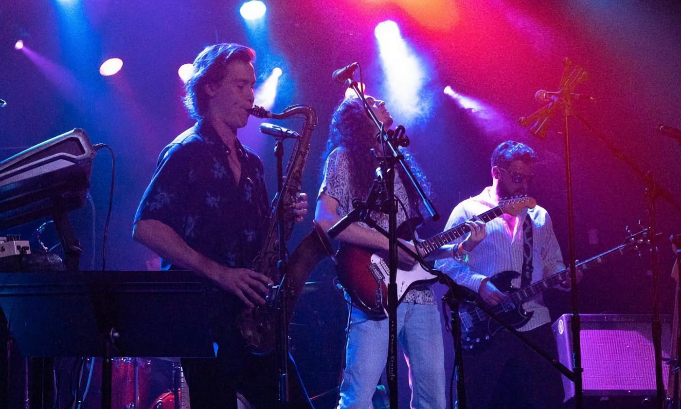 Three musicians from the band Star Collar (saxophone, bass, guitar) on a stage beneath multi color lights.
