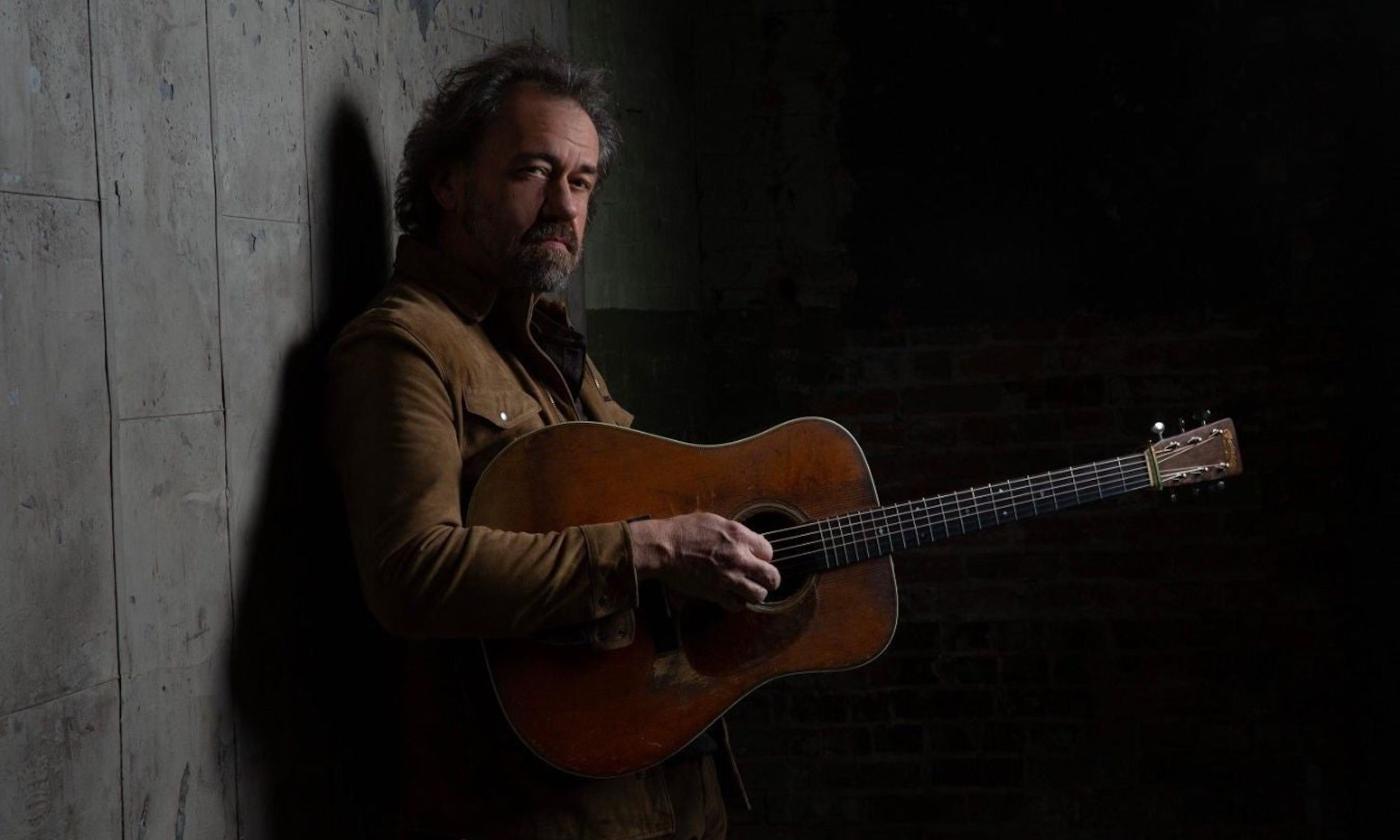 Dan Tyminski leans against a gray wall and strums his guitar.