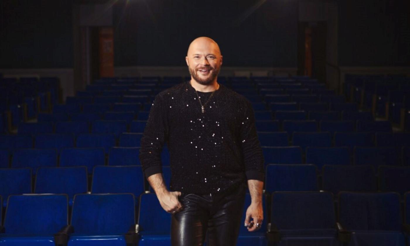 Travis Holp wears black clothing while posing in front of a row of blue chairs. 