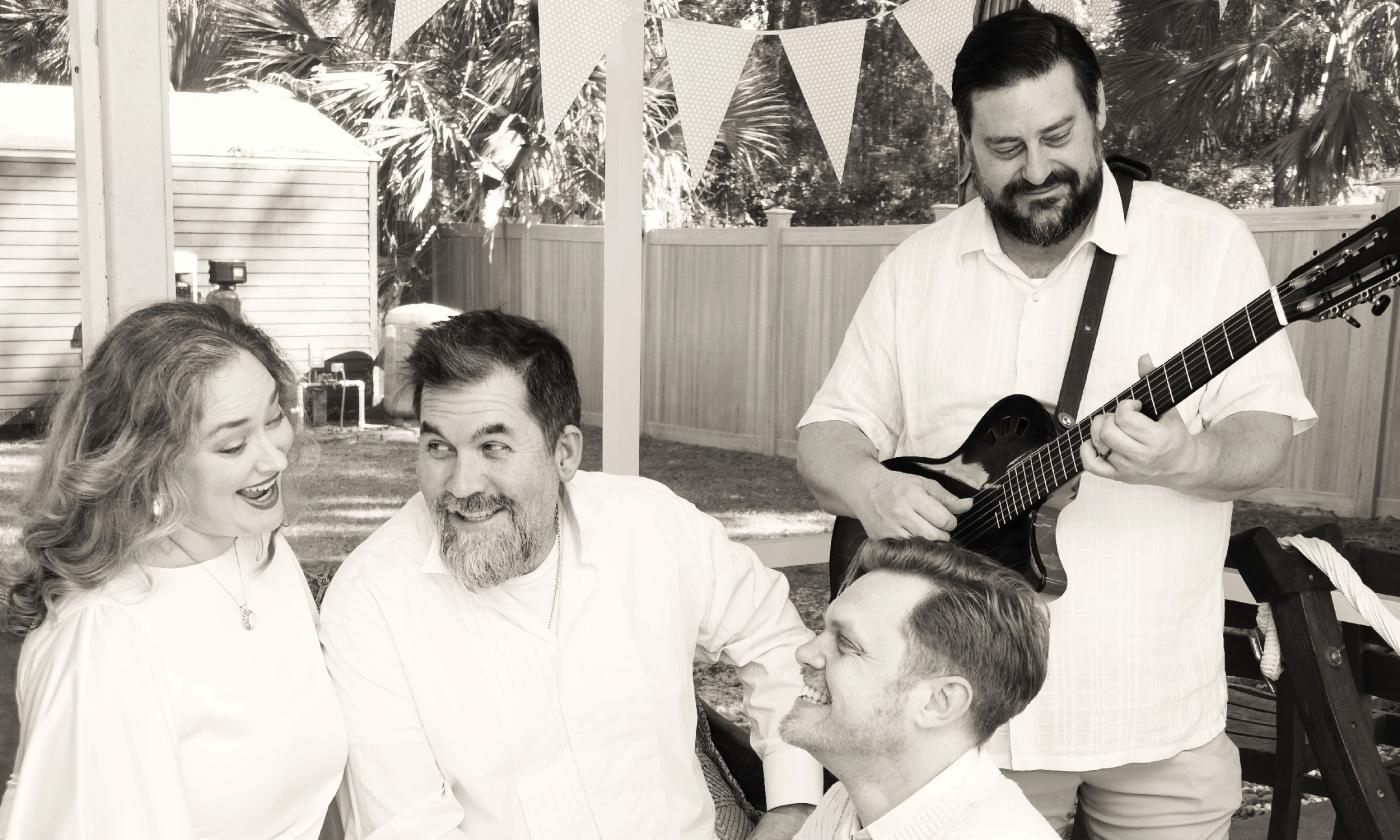 In black and white, the four members of the Pinnacle Singers, wearing white, one with a guitar