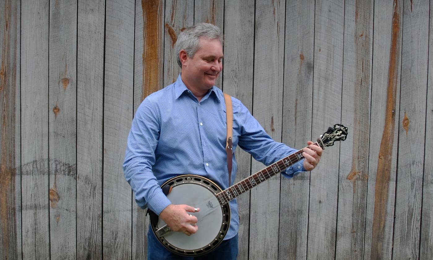 Scott Anderson, wearing a blue shirt and playing banjo in front of a grey board fence