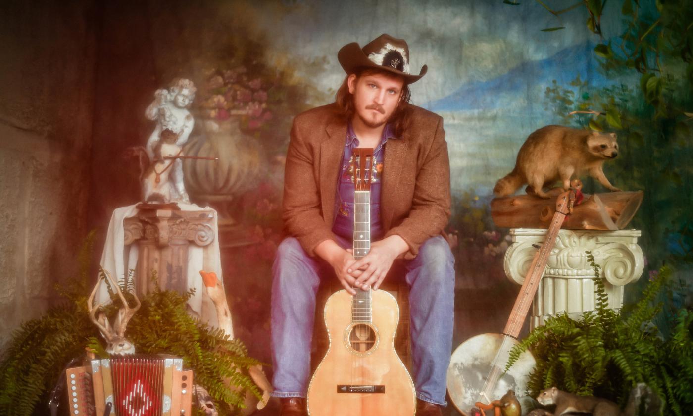 Musician and songwriter Willi Carlisle, seated with props and a colorful backdrop