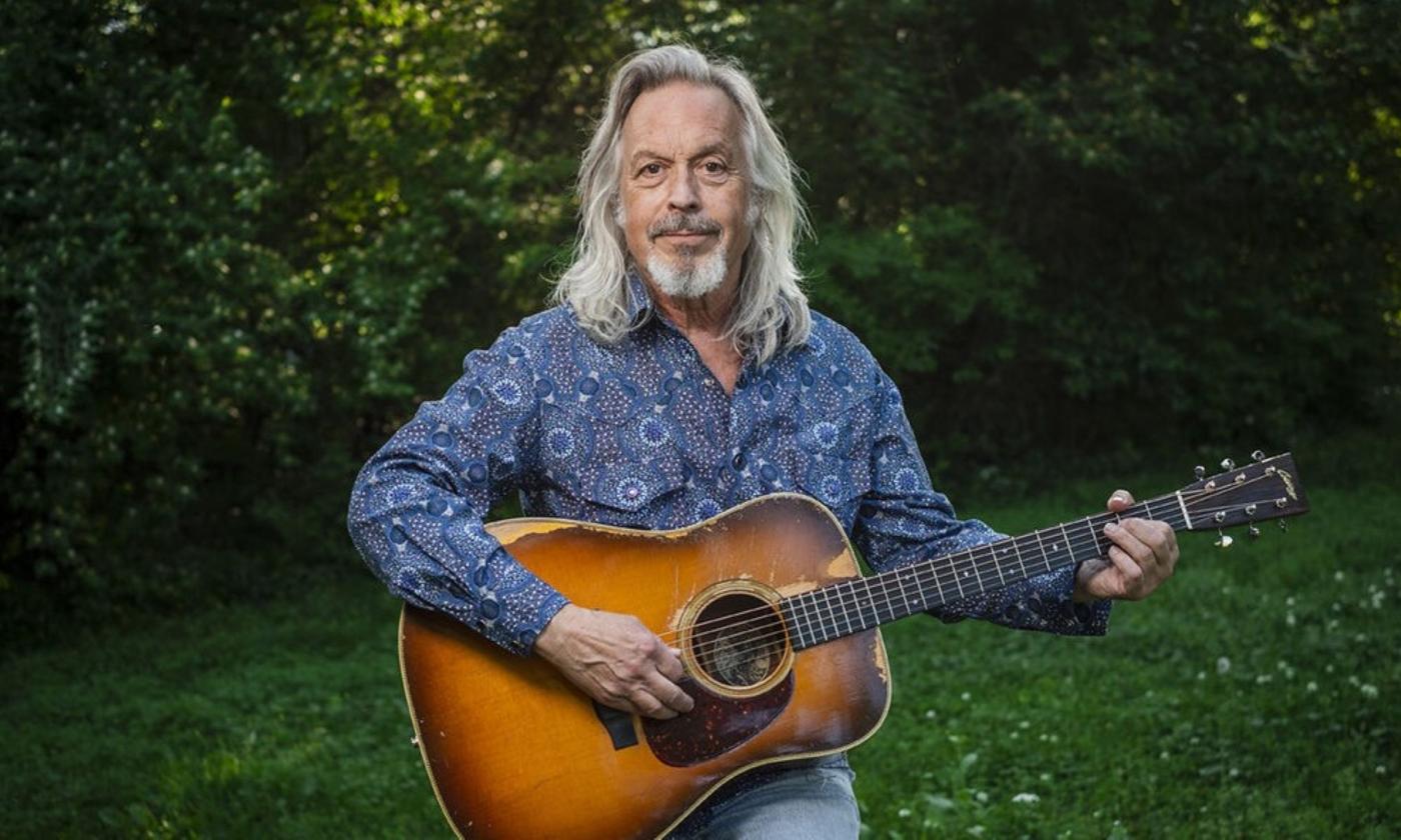 Jim Lauderdale poses with his guitar against a backdrop of lush green foliage.