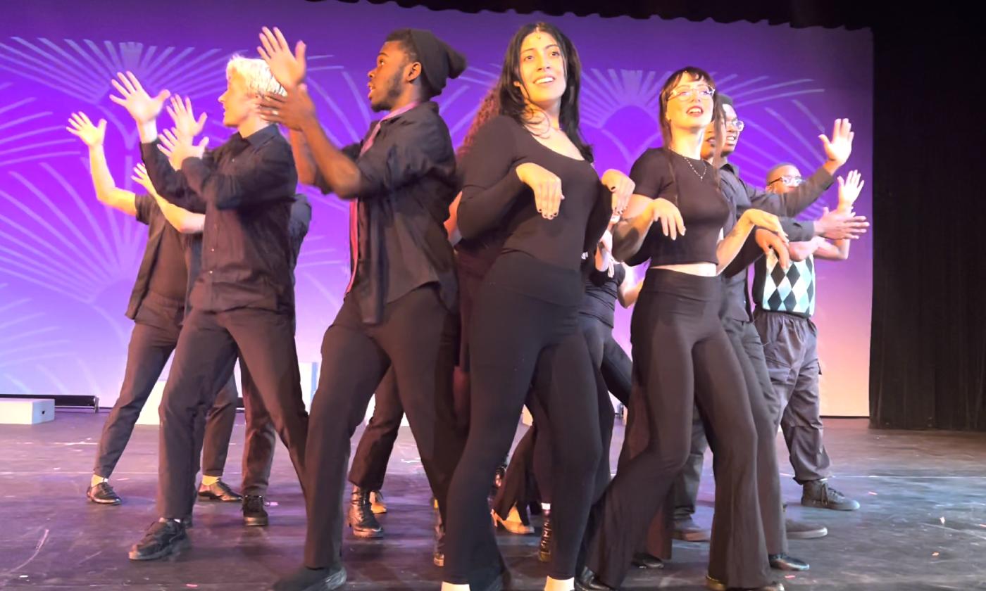 Dancers in dark clothing perform a broadway tune on stage with a purple background