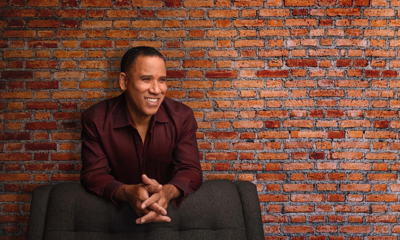 A singer, leans on a grey chair while standing in front of a brick wall