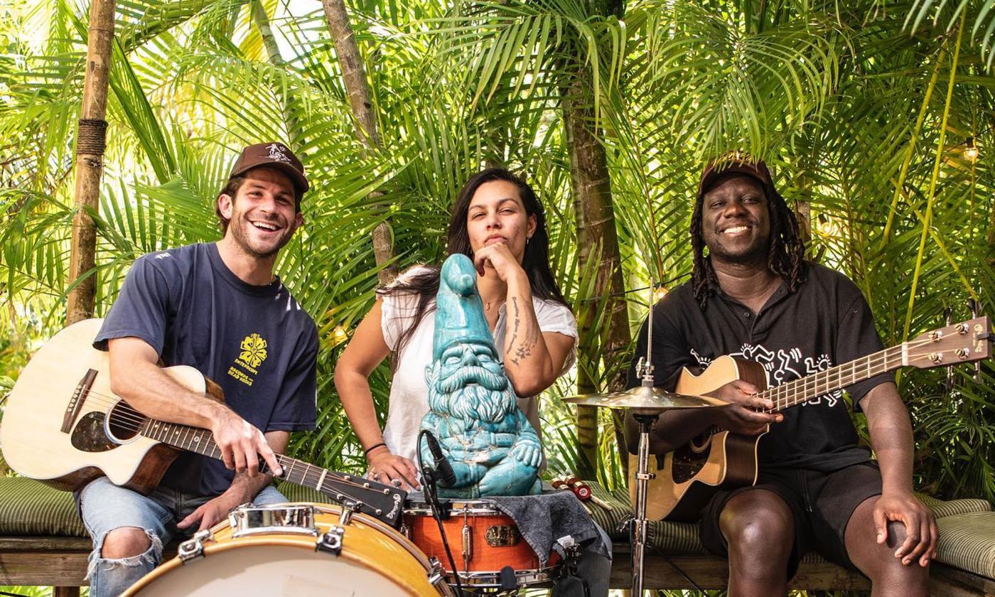 The band Surfer Girl sitting outside in tropical setting, with instruments, all smiling
