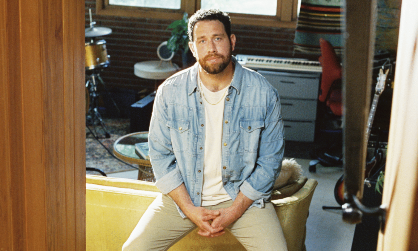 Jordan Fletcher strikes a relaxed pose on the edge of a couch, wearing a classic denim shirt.