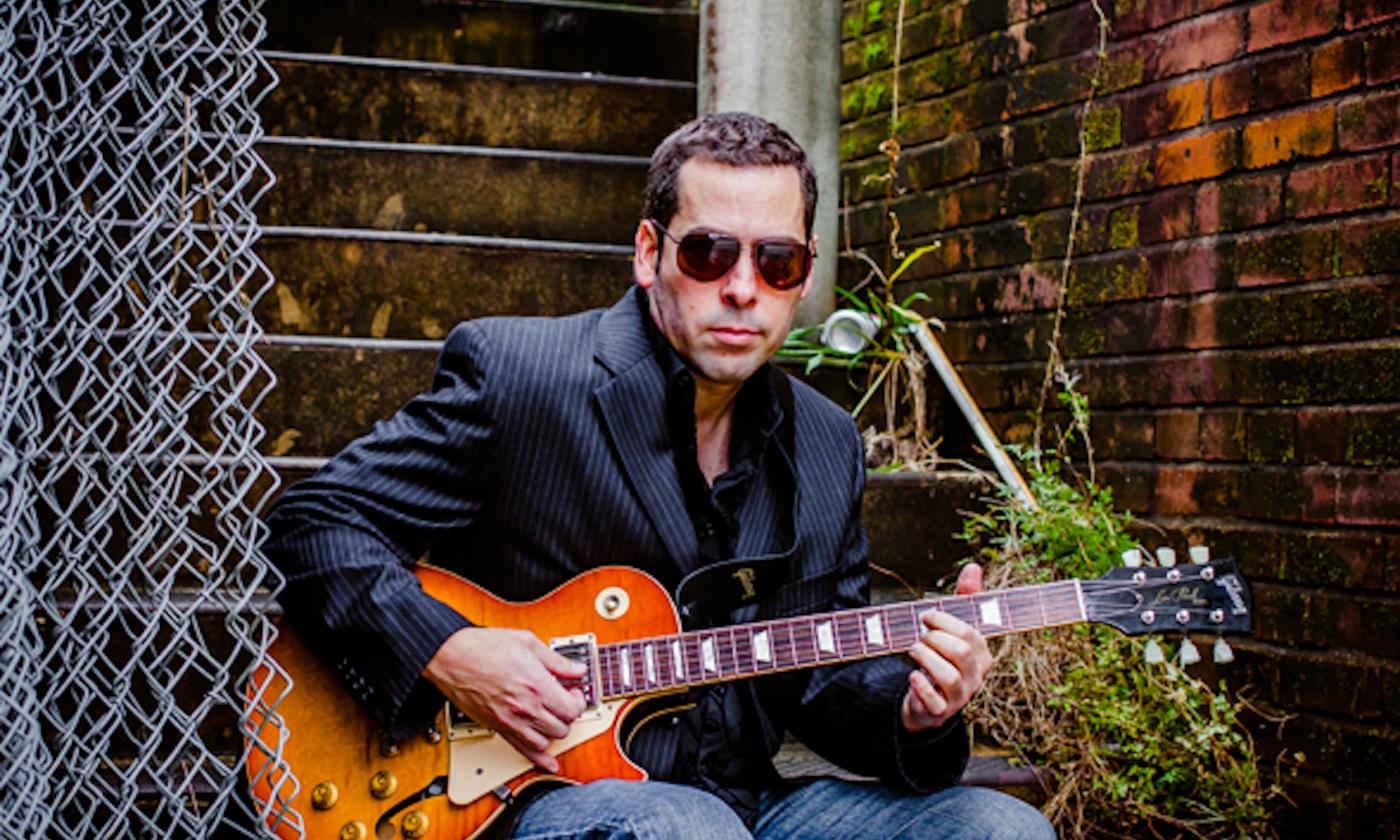 Albert Castiglia wears glasses while strumming his guitar.