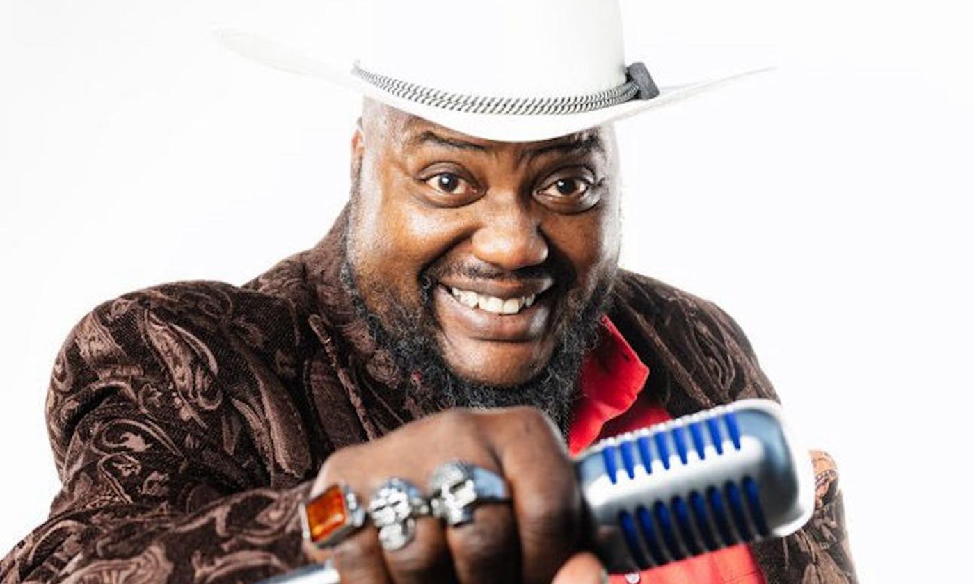 Sugar Rayford smiles while clutching a mic in front of a white backdrop.