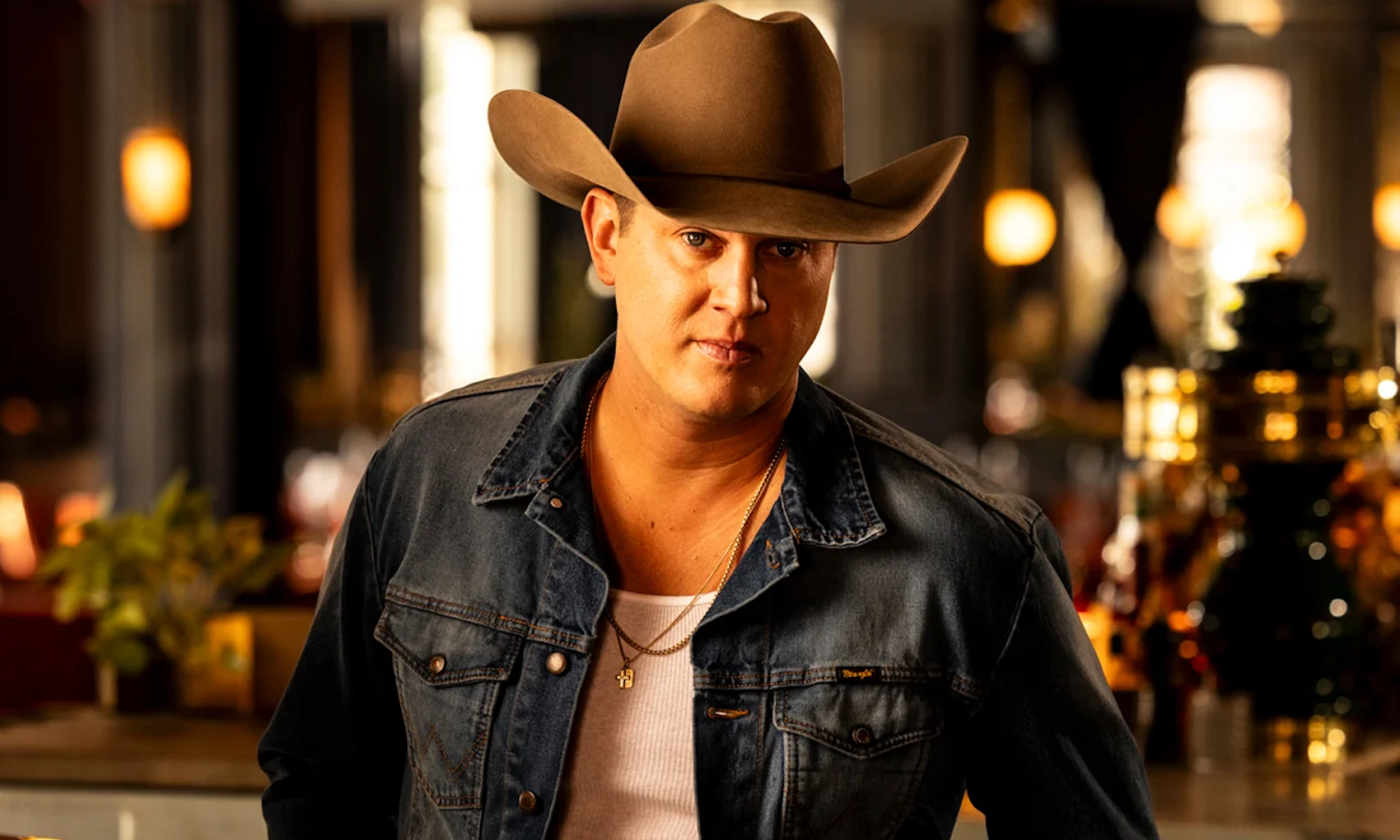 Jon Pardi poses with a brown cowboy hat and dark blue denim jacket in front of a bar with brown and amber hues.