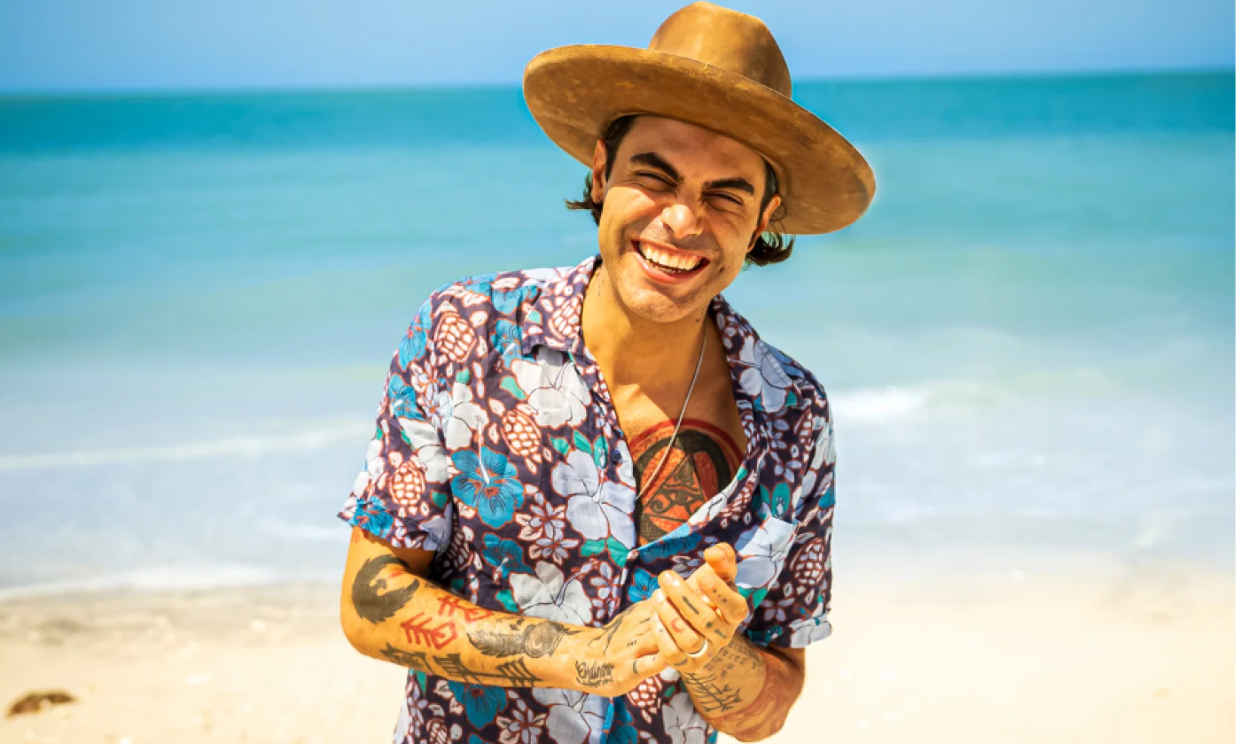 Niko Moon clasps his hands together and stands in front of a sandy beach.