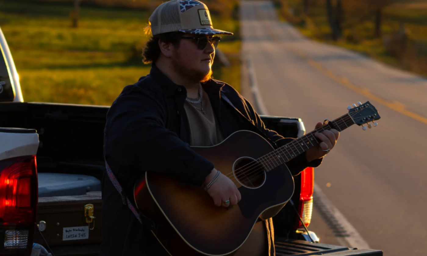 Bayker Blankenship leans on his truck and strums his guitar.