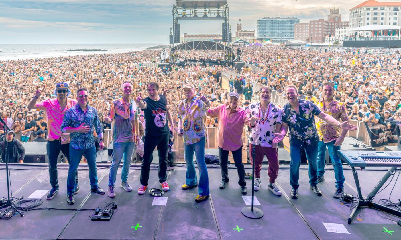 The Beach Boys smile and pose on stage with a lively concert crowd behind them.