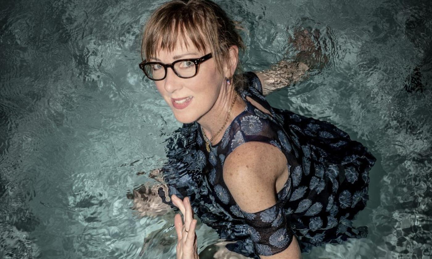 Jonatha Brooke, wearing glasses, smiles warmly as she poses against a textured background.