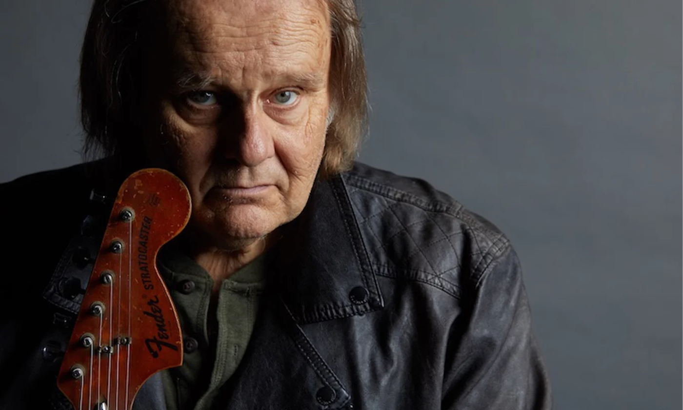 Walter Trout clutches his guitar and poses in front of a gray backdrop.