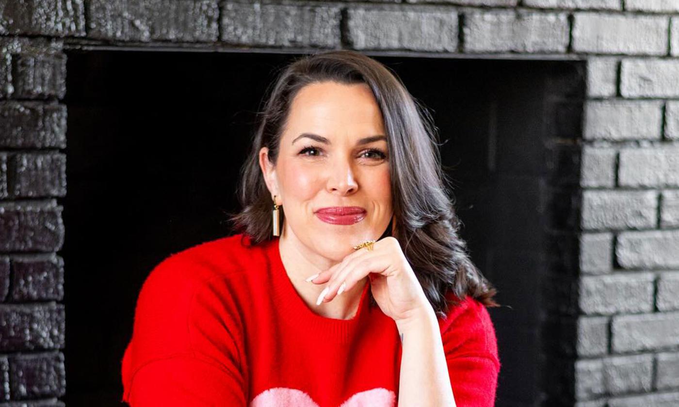 Songwriter Amy Gerhartz in front of a fire place, smiling, with her hand to her cheek