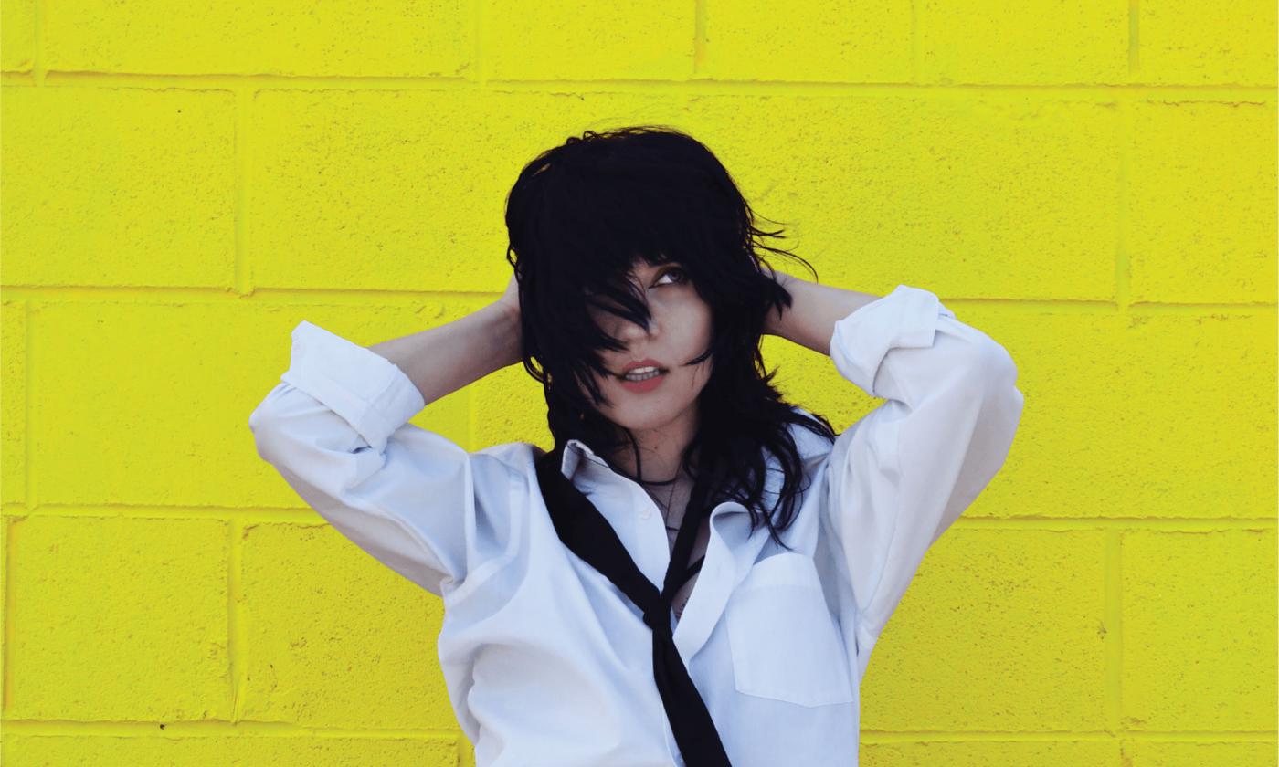 Patty PerShayla standing in front of chartreuse wall, in white blouse and black tie. Image by Rae Jean Erickson