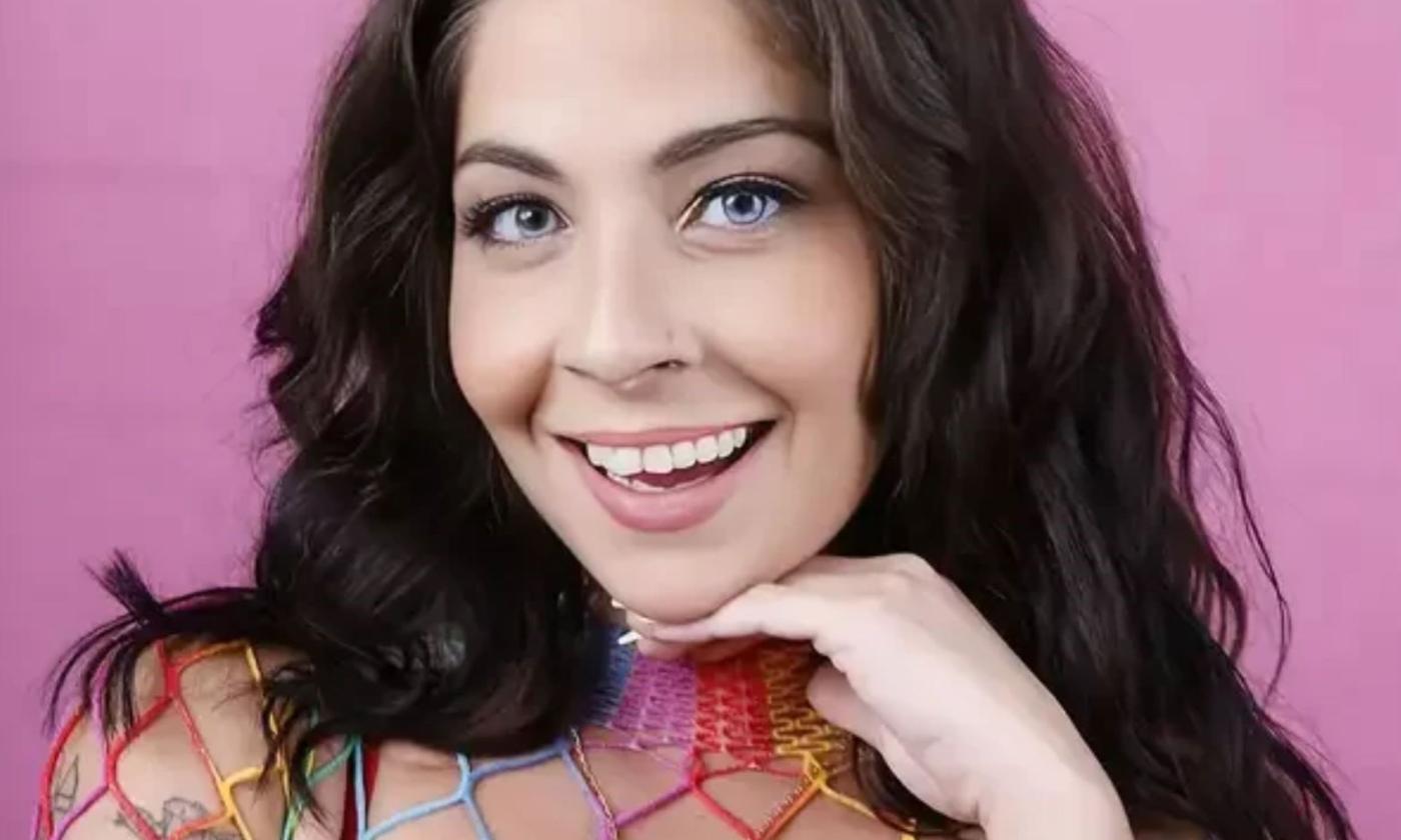 Comedian Brittany Brave smiles while posing in front of a pink backdrop.