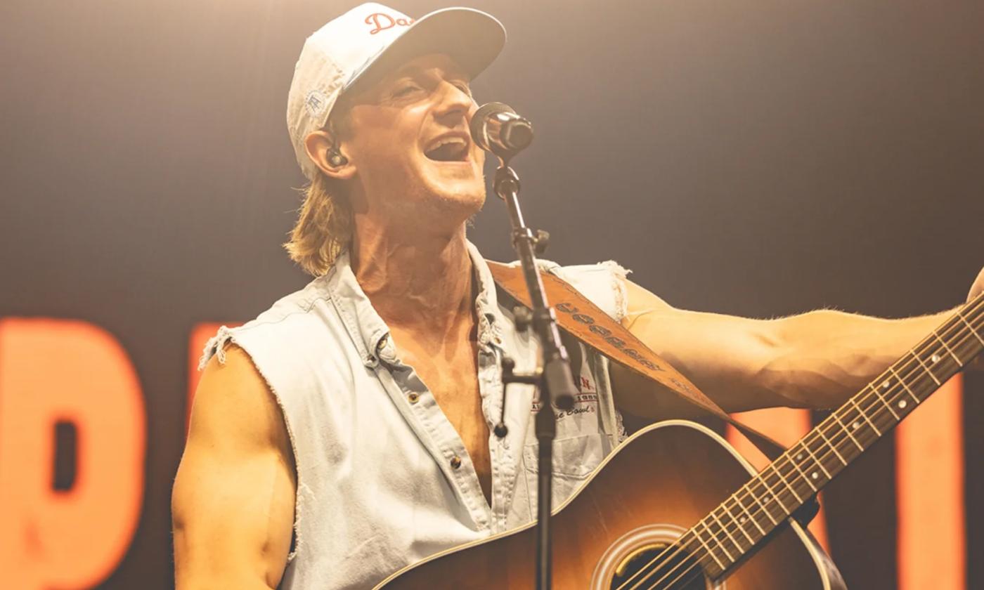 Cooper Alan wears a hat and strums his guitar on stage. 