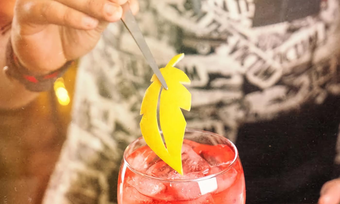 The hand of a bartender adding an artistic cut lemon skin to a pink cocktail