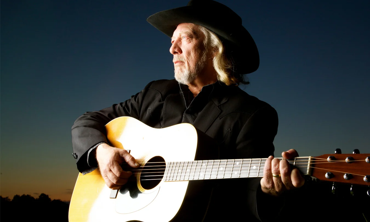 John Anderson dons a black cowboy hat and strums his guitar. 